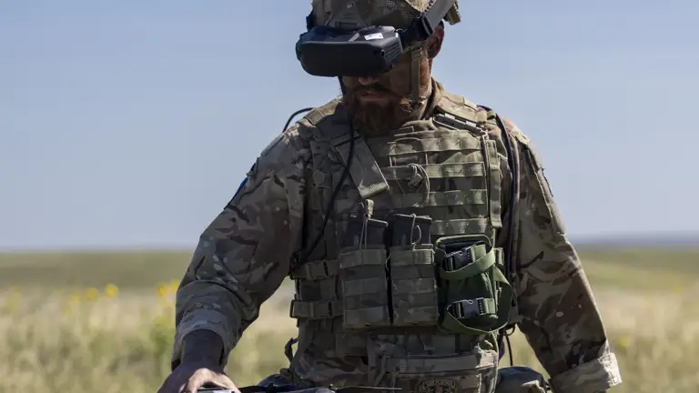 A person in camouflage military gear stands in an open grassy field, holding a drone. The person is wearing a helmet and protective eyewear, with various equipment attached to their vest. The sky is clear and blue.