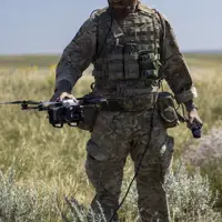 A person in camouflage military gear stands in an open grassy field, holding a drone. The person is wearing a helmet and protective eyewear, with various equipment attached to their vest. The sky is clear and blue.