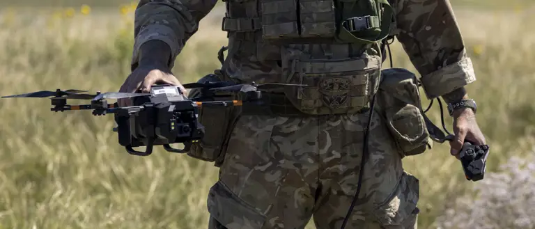 A person in camouflage military gear stands in an open grassy field, holding a drone. The person is wearing a helmet and protective eyewear, with various equipment attached to their vest. The sky is clear and blue.