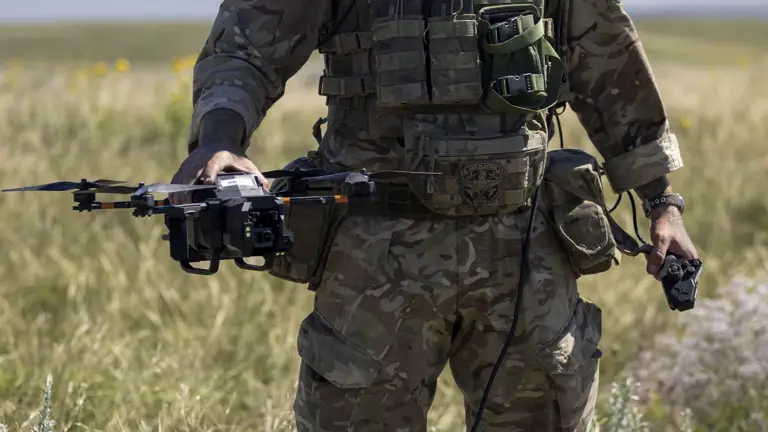 A person in camouflage military gear stands in an open grassy field, holding a drone. The person is wearing a helmet and protective eyewear, with various equipment attached to their vest. The sky is clear and blue.