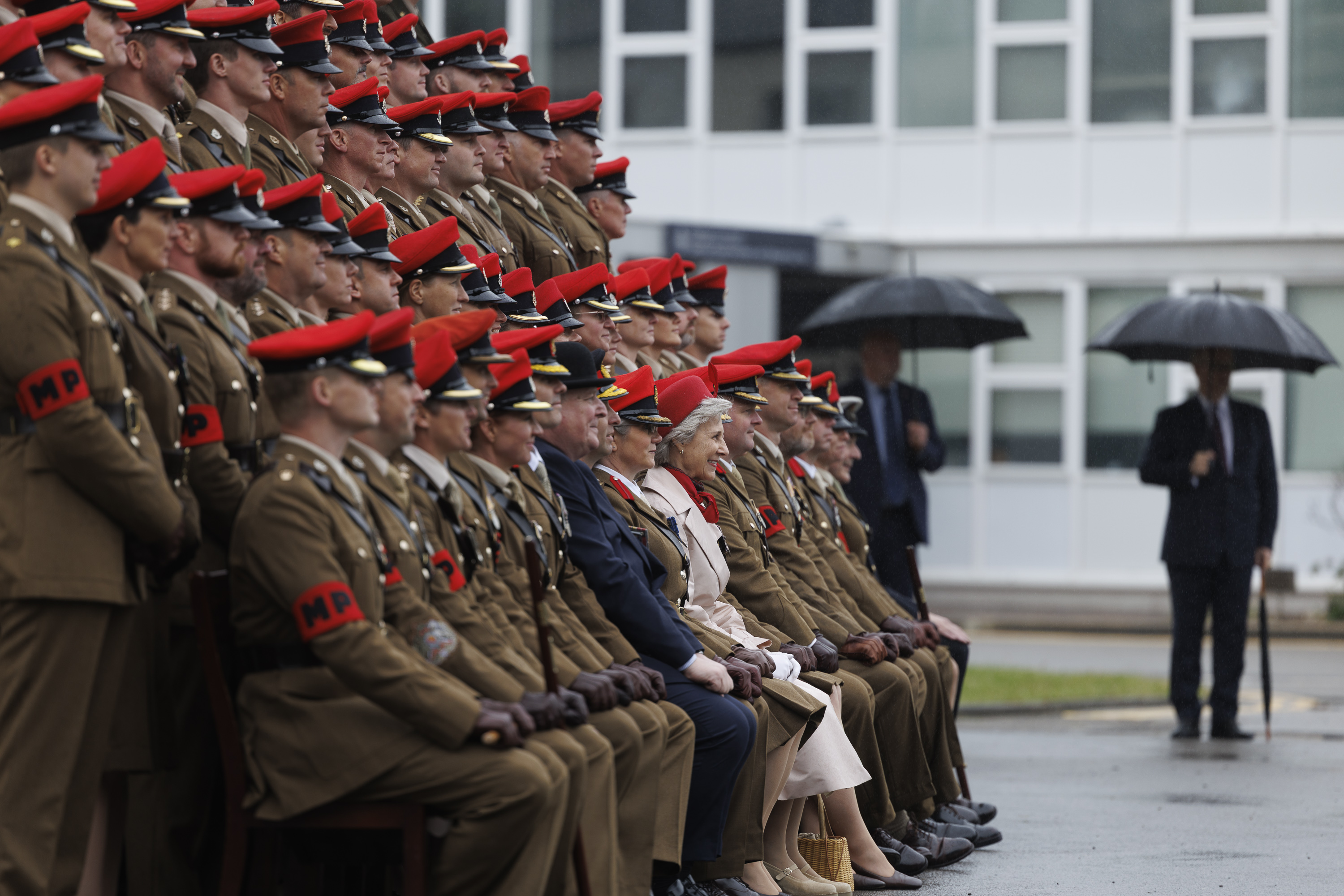 Soldiers sat in line next to each other wearing their brown smart uniforms with red caps on their heads. A women wearing a cream coat and a red hat is sat in the middle at the front. Two men in blue suits are seen next to them.