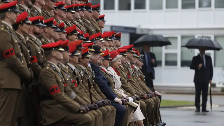 Soldiers sat in line next to each other wearing their brown smart uniforms with red caps on their heads. A women wearing a cream coat and a red hat is sat in the middle at the front. Two men in blue suits are seen next to them.