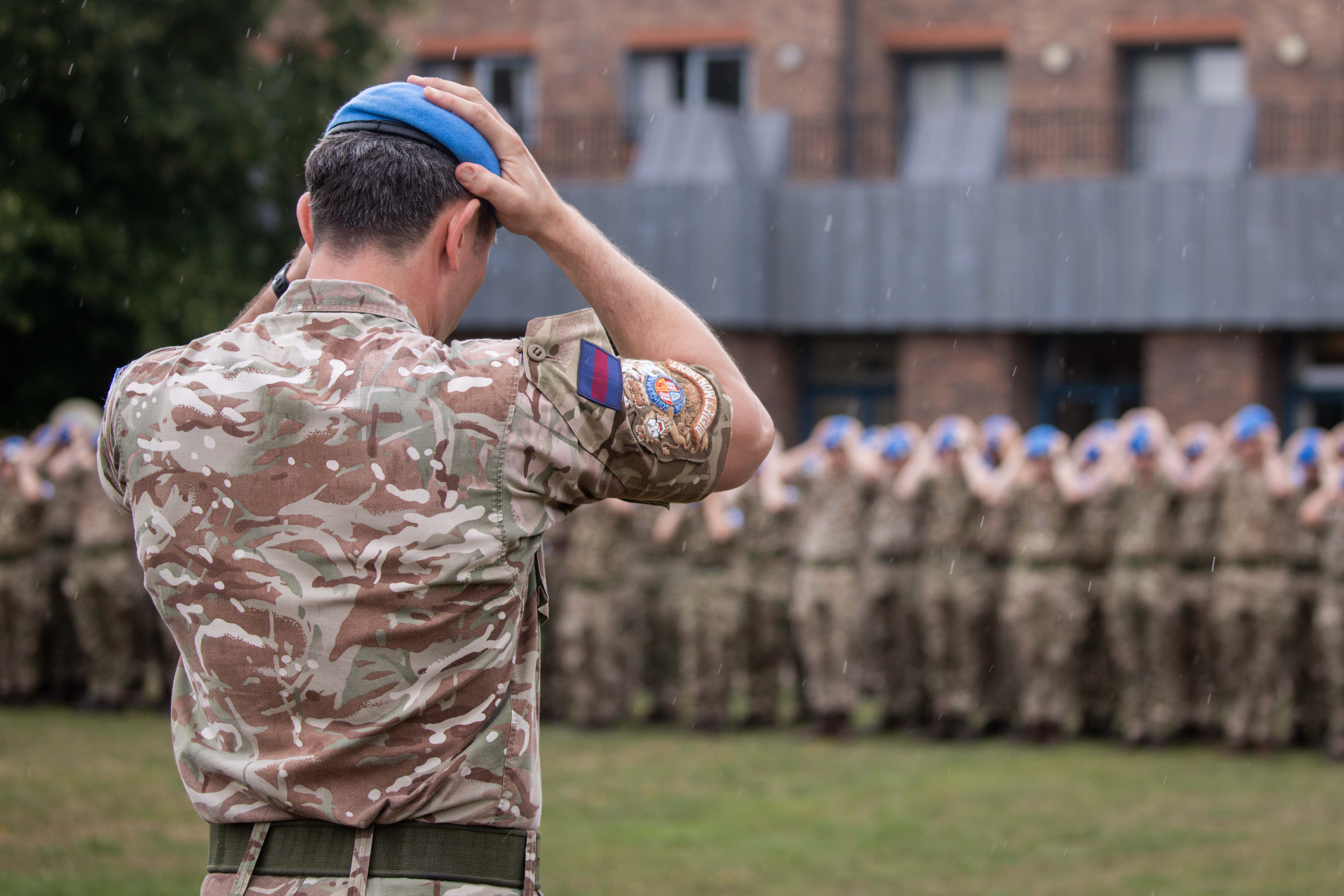 Soldier in camouflage uniform adjusting blue beret while facing a formation of fellow soldiers.
