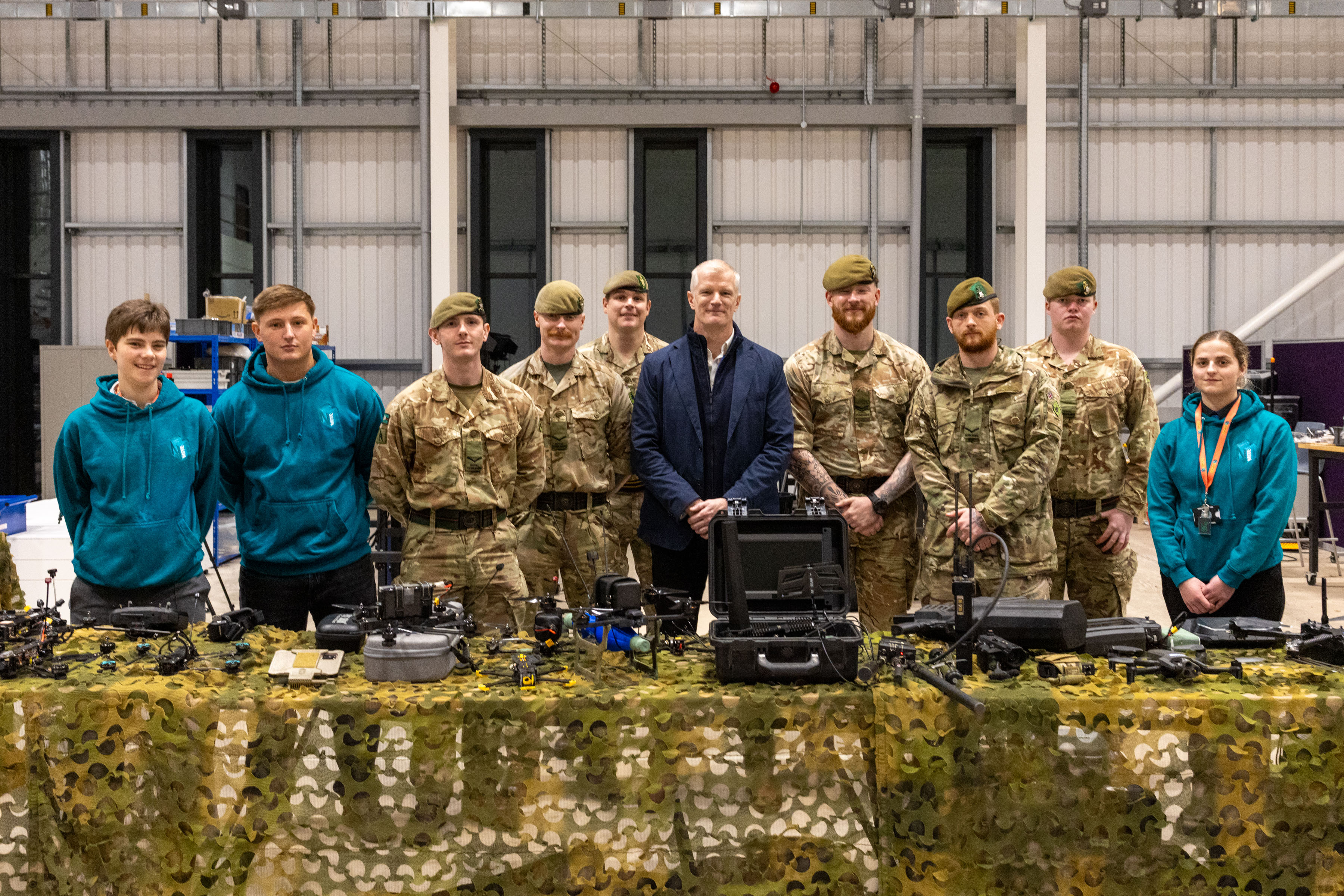 Group of military personnel and civilians standing behind a table with various equipment in an industrial setting.