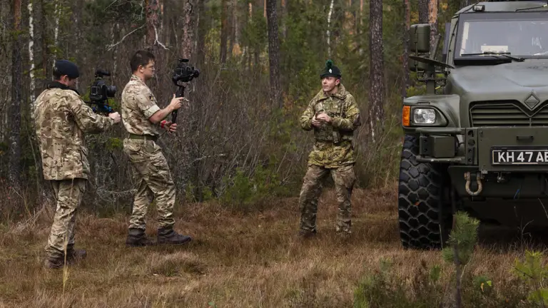 Two Army Photographers stand in a wooded area with a military vehicle off to the right hand side. They are filming and interviewing a soldier from an Irish regiment.