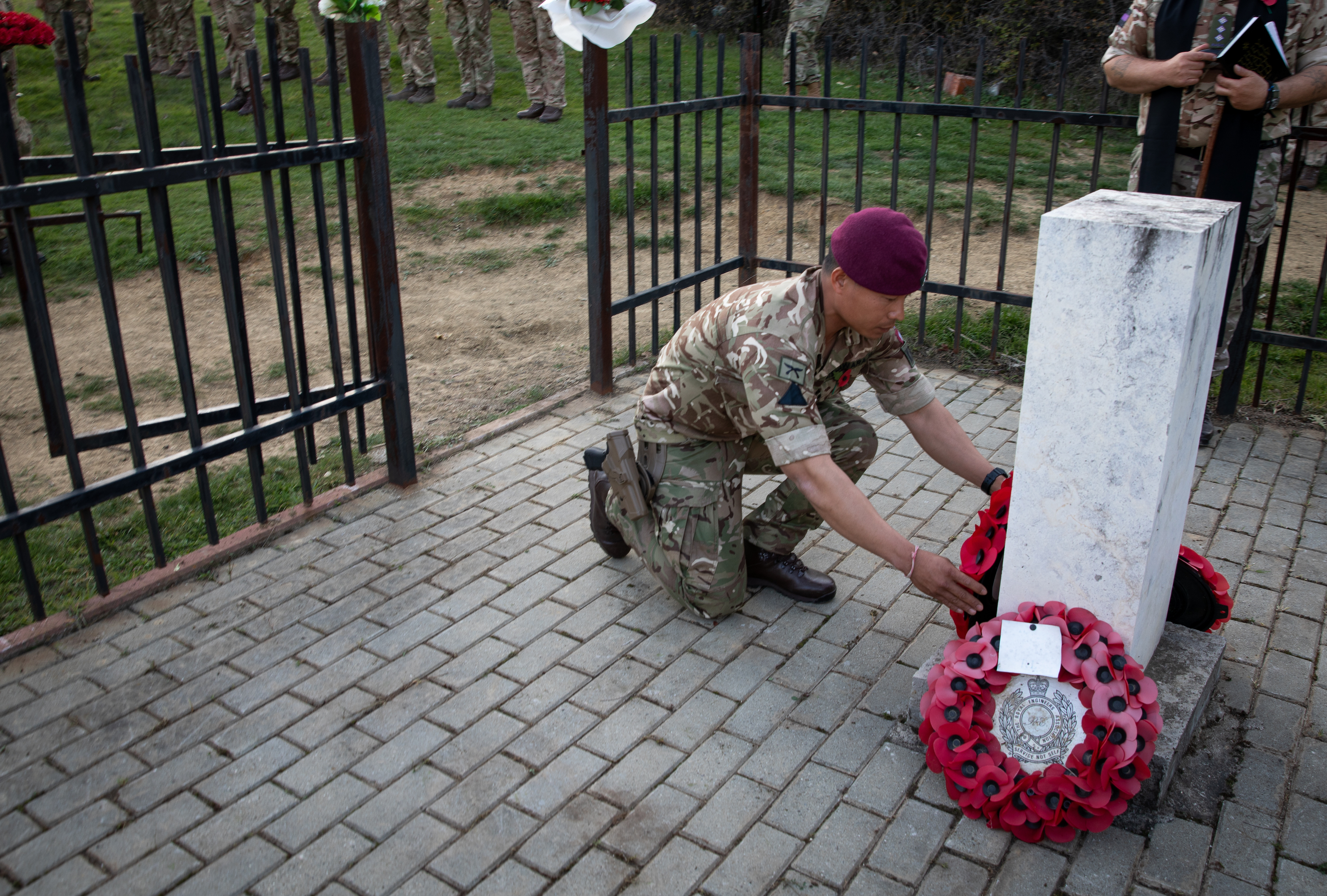 A soldier wearing camouflage uniform and a maroon beret kneels down to lay a poppy wreath.