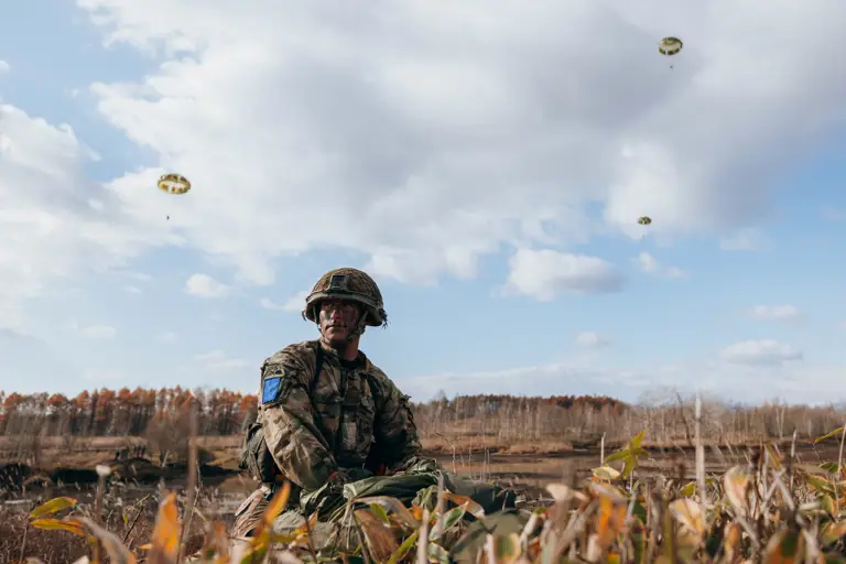Soldier in camouflage gear sitting in a field with parachutes descending in the cloudy sky above.
