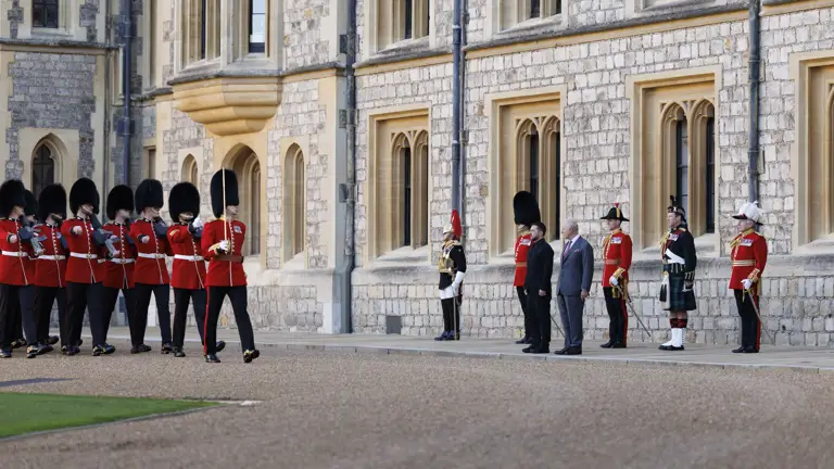 British guards in red uniforms and bearskin hats marching past officials standing outside of a historic stone building.