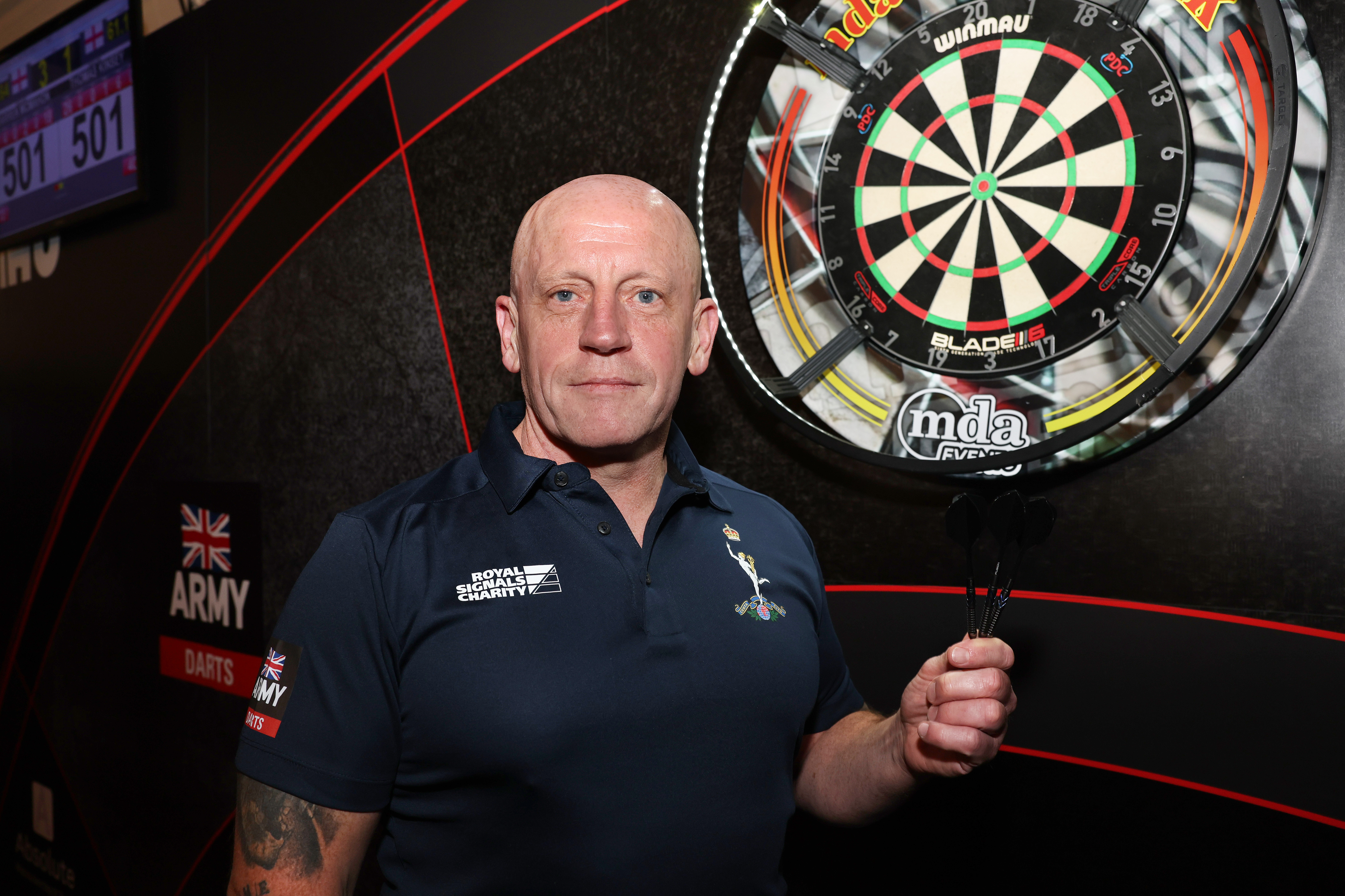 Man in a navy blue Royal Signals shirt holding  darts in a darts competition setting.