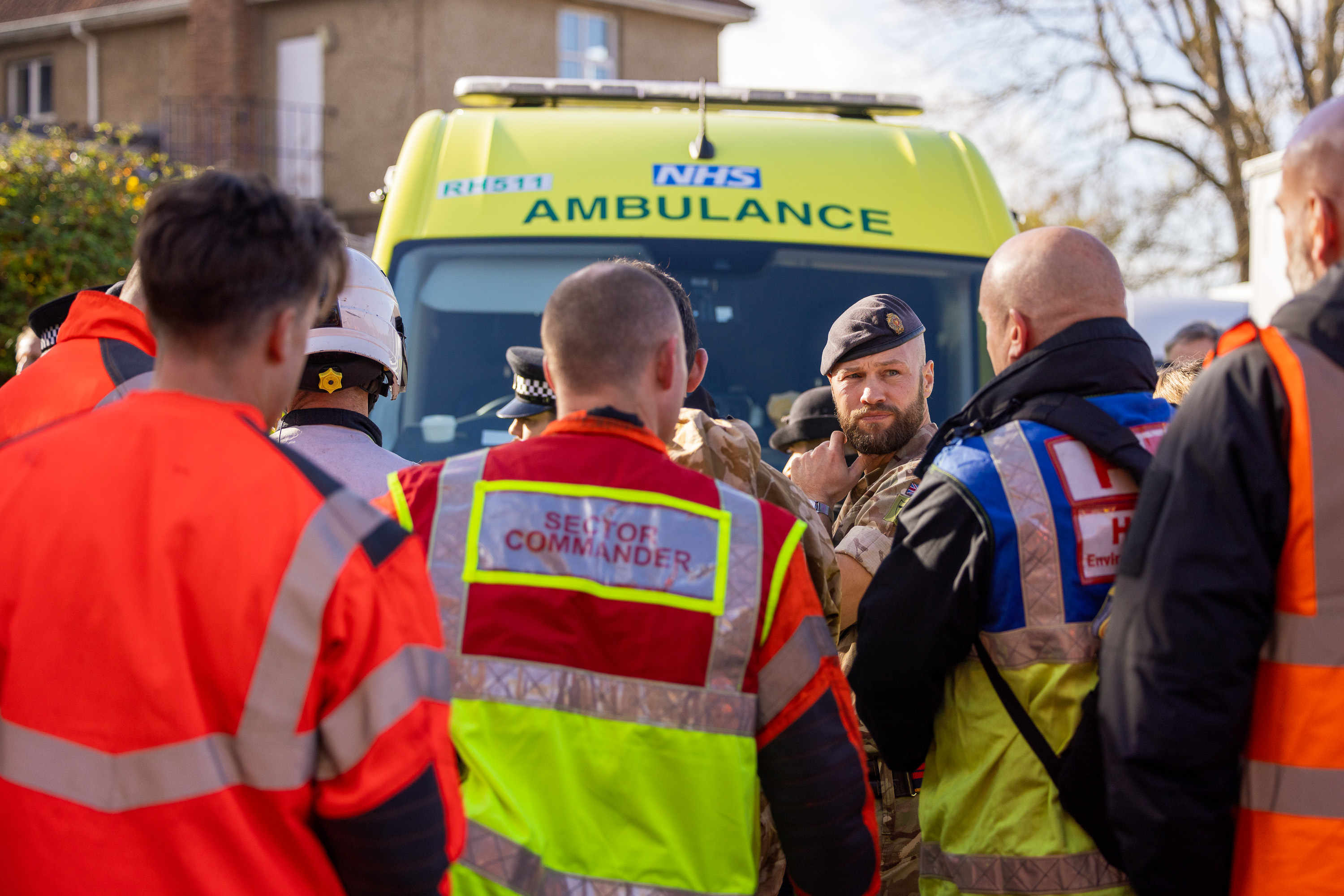 Group of emergency responders in high-visibility jackets gathered near a yellow NHS ambulance during a coordinated operation.