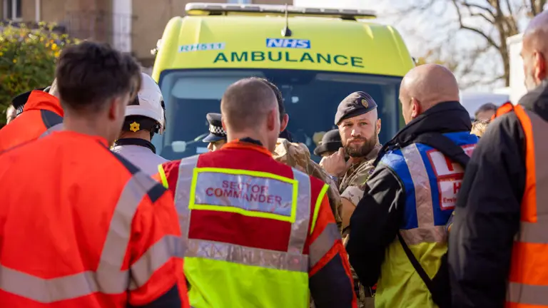Group of emergency responders in high-visibility jackets gathered near a yellow NHS ambulance during a coordinated operation.