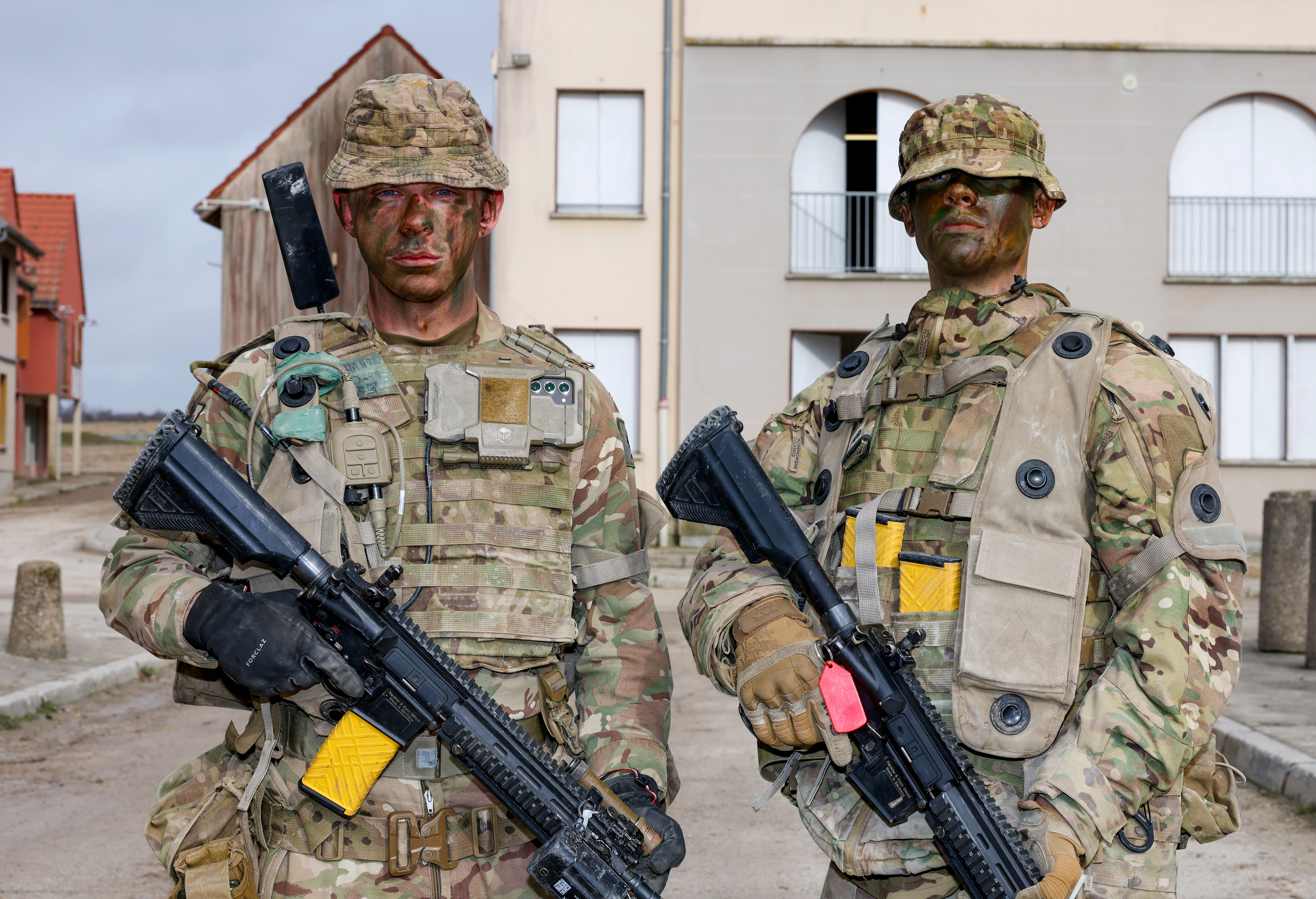Two individuals dressed in full military camouflage gear stand in an urban environment, holding rifles. 