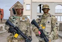 Two individuals dressed in full military camouflage gear stand in an urban environment, holding rifles.