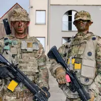 Two individuals dressed in full military camouflage gear stand in an urban environment, holding rifles.