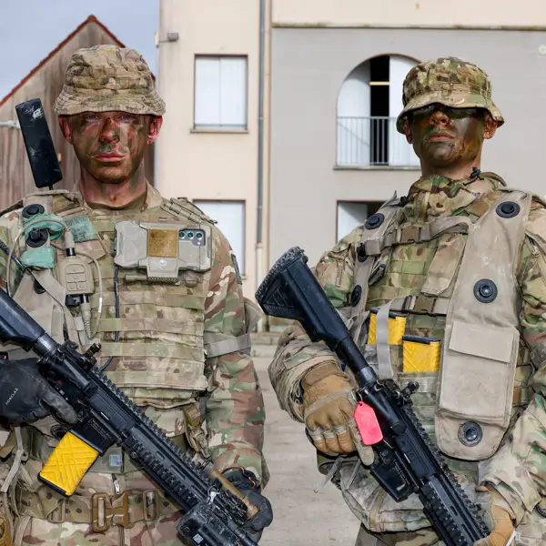 Two individuals dressed in full military camouflage gear stand in an urban environment, holding rifles.