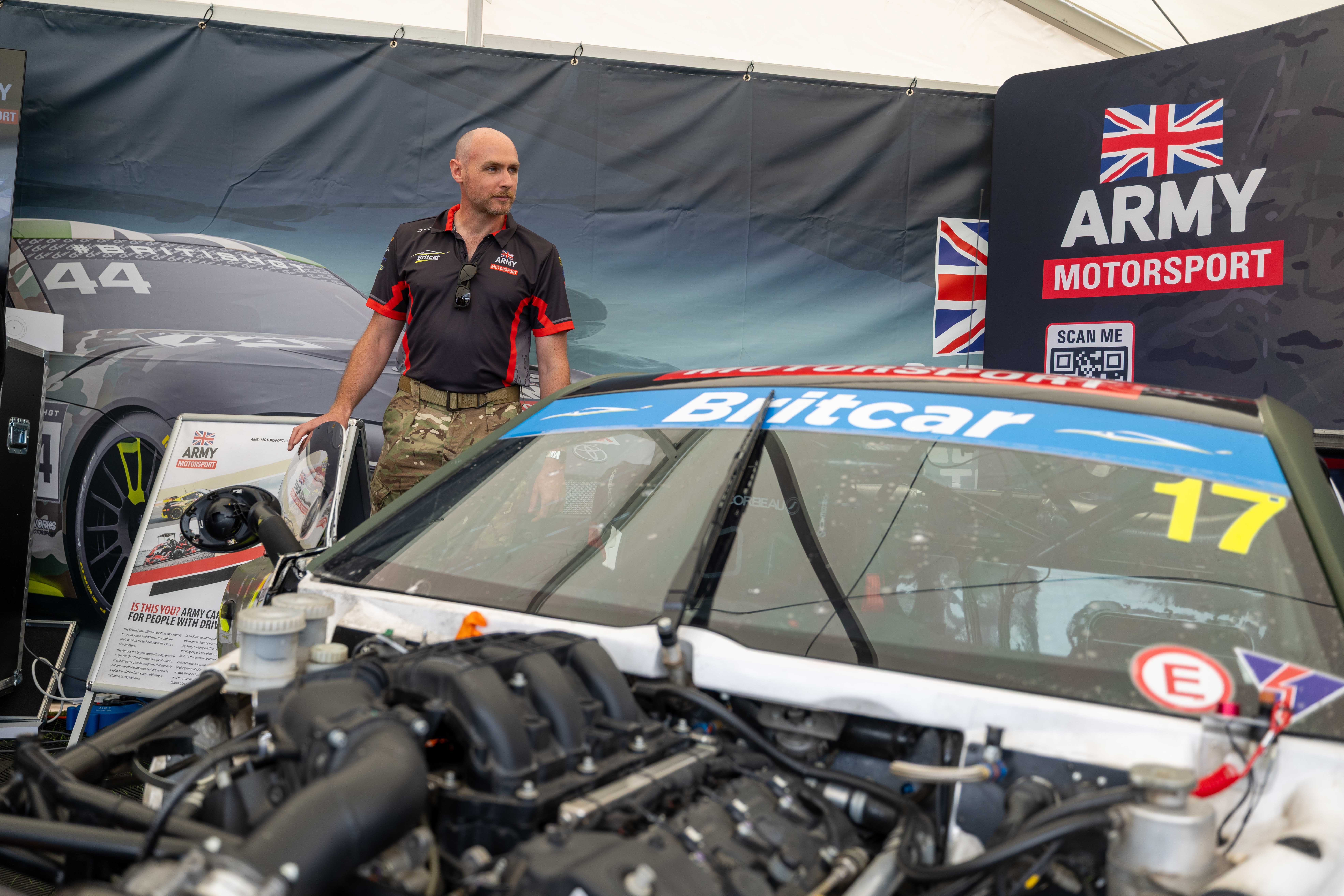 A person in army gear stands beside a racing car labeled 