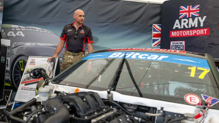A person in army gear stands beside a racing car labeled