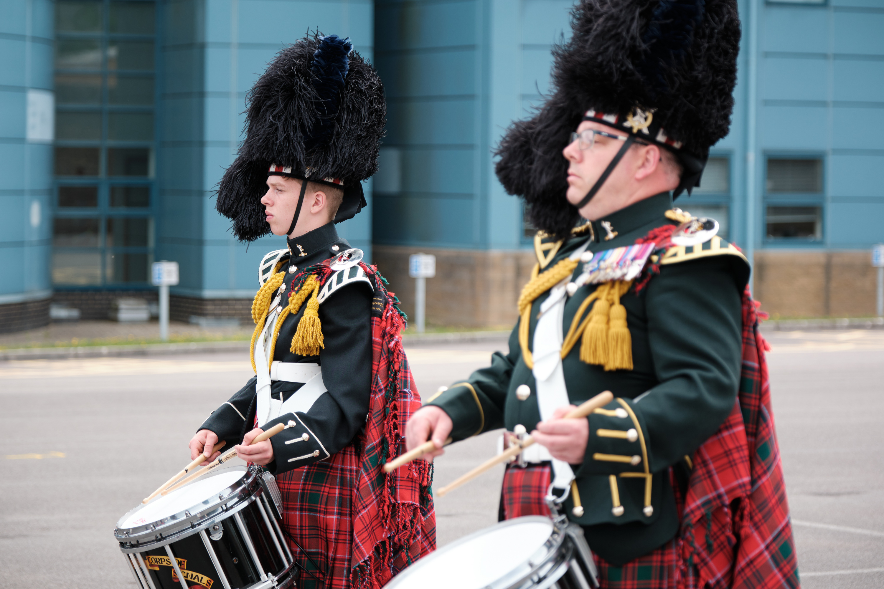 Two drummers in traditional Scottish military uniforms with feathered hats and tartan kilts performing outdoors.