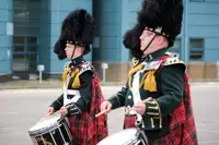 Two drummers in traditional Scottish military uniforms with feathered hats and tartan kilts performing outdoors.
