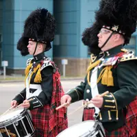 Two drummers in traditional Scottish military uniforms with feathered hats and tartan kilts performing outdoors.