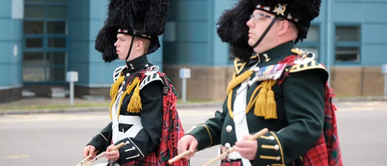Two drummers in traditional Scottish military uniforms with feathered hats and tartan kilts performing outdoors.