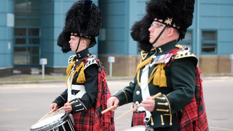 Two drummers in traditional Scottish military uniforms with feathered hats and tartan kilts performing outdoors.