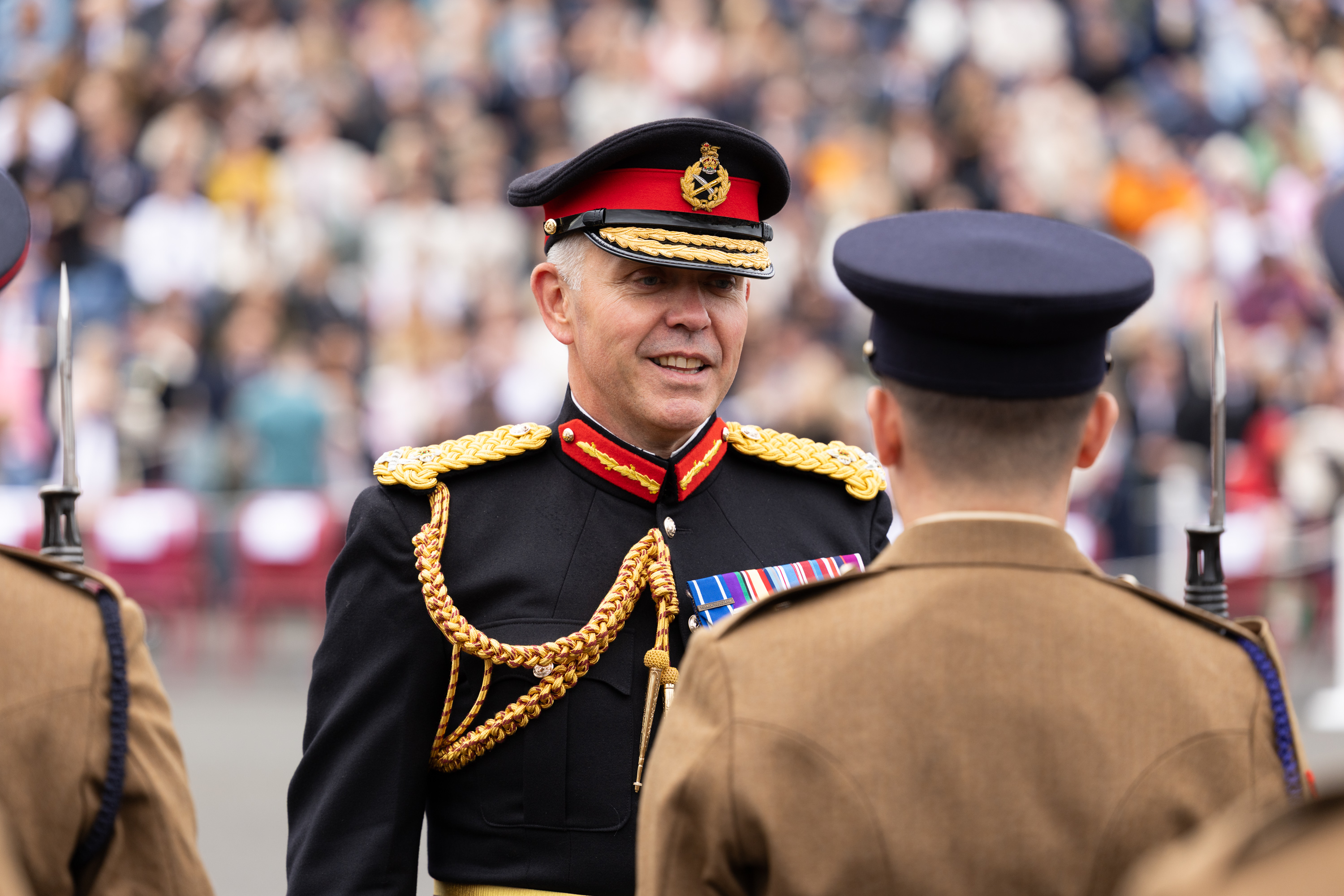 A Senior Army Officer in a dark blue uniform talks with a soldier wearing a khaki uniform. In the background seated in raised stands, are the families of the soldiers