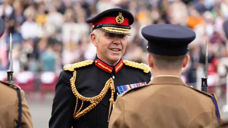 A Senior Army Officer in a dark blue uniform talks with a soldier wearing a khaki uniform. In the background seated in raised stands, are the families of the soldiers