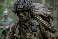 Soldier wearing camouflage uniform and helmet and body armour seen in the field, a bag on his back and a string of general purpose machine gun ammunition going around his body.