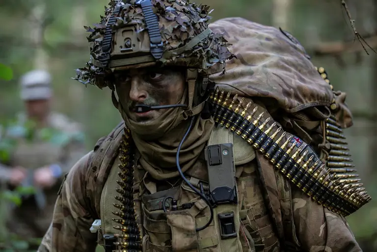 Soldier wearing camouflage uniform and helmet and body armour seen in the field, a bag on his back and a string of general purpose machine gun ammunition going around his body.