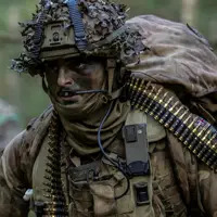 Soldier wearing camouflage uniform and helmet and body armour seen in the field, a bag on his back and a string of general purpose machine gun ammunition going around his body.