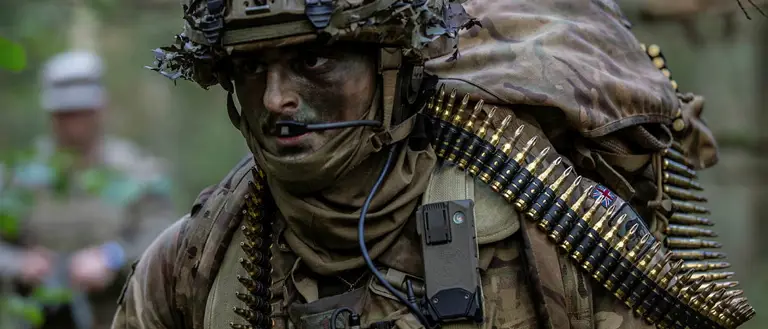 Soldier wearing camouflage uniform and helmet and body armour seen in the field, a bag on his back and a string of general purpose machine gun ammunition going around his body.