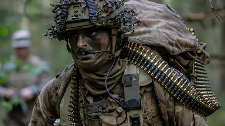 Soldier wearing camouflage uniform and helmet and body armour seen in the field, a bag on his back and a string of general purpose machine gun ammunition going around his body.
