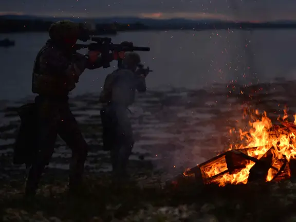 Two soldiers in the dark by a body of water with a roaring fire next to them. The soldiers are holding guns.