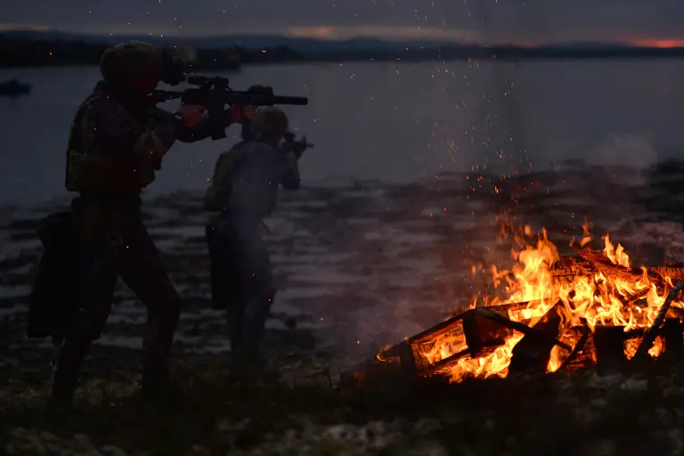 Two soldiers in the dark by a body of water with a roaring fire next to them. The soldiers are holding guns.