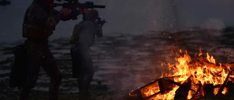 Two soldiers in the dark by a body of water with a roaring fire next to them. The soldiers are holding guns.