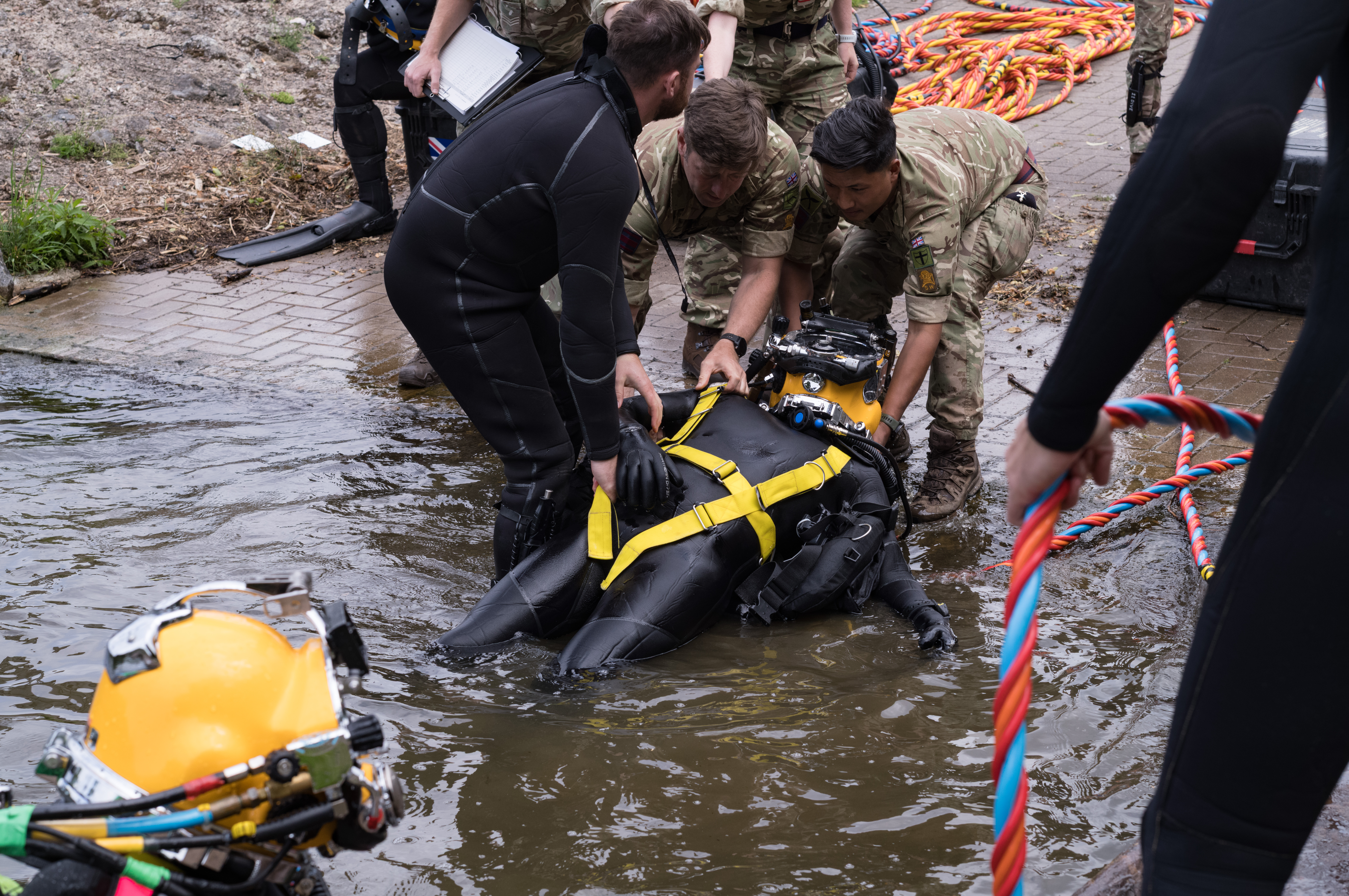 Military personnel assist a diver in a black wetsuit with yellow harness as they emerge from water near a paved area. 
