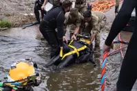Military personnel assist a diver in a black wetsuit with yellow harness as they emerge from water near a paved area.