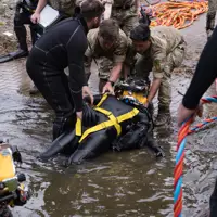 Military personnel assist a diver in a black wetsuit with yellow harness as they emerge from water near a paved area.