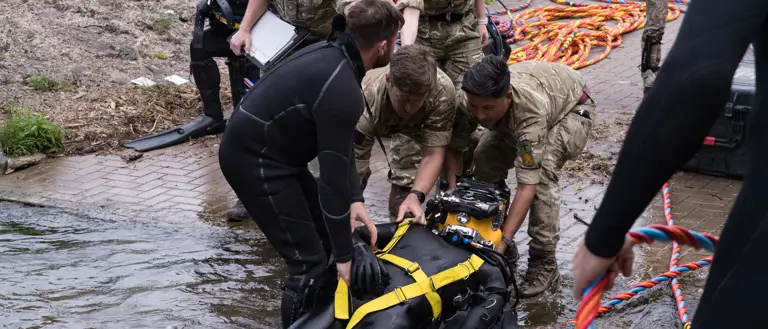 Military personnel assist a diver in a black wetsuit with yellow harness as they emerge from water near a paved area.