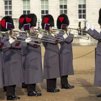 British guards in traditional bearskin hats and grey coats playing trombones in formation during a ceremonial event.