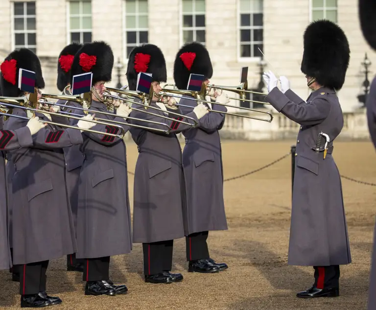 British guards in traditional bearskin hats and grey coats playing trombones in formation during a ceremonial event.