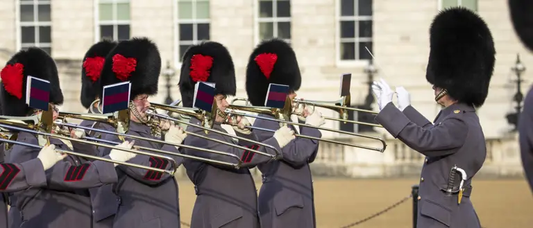 British guards in traditional bearskin hats and grey coats playing trombones in formation during a ceremonial event.
