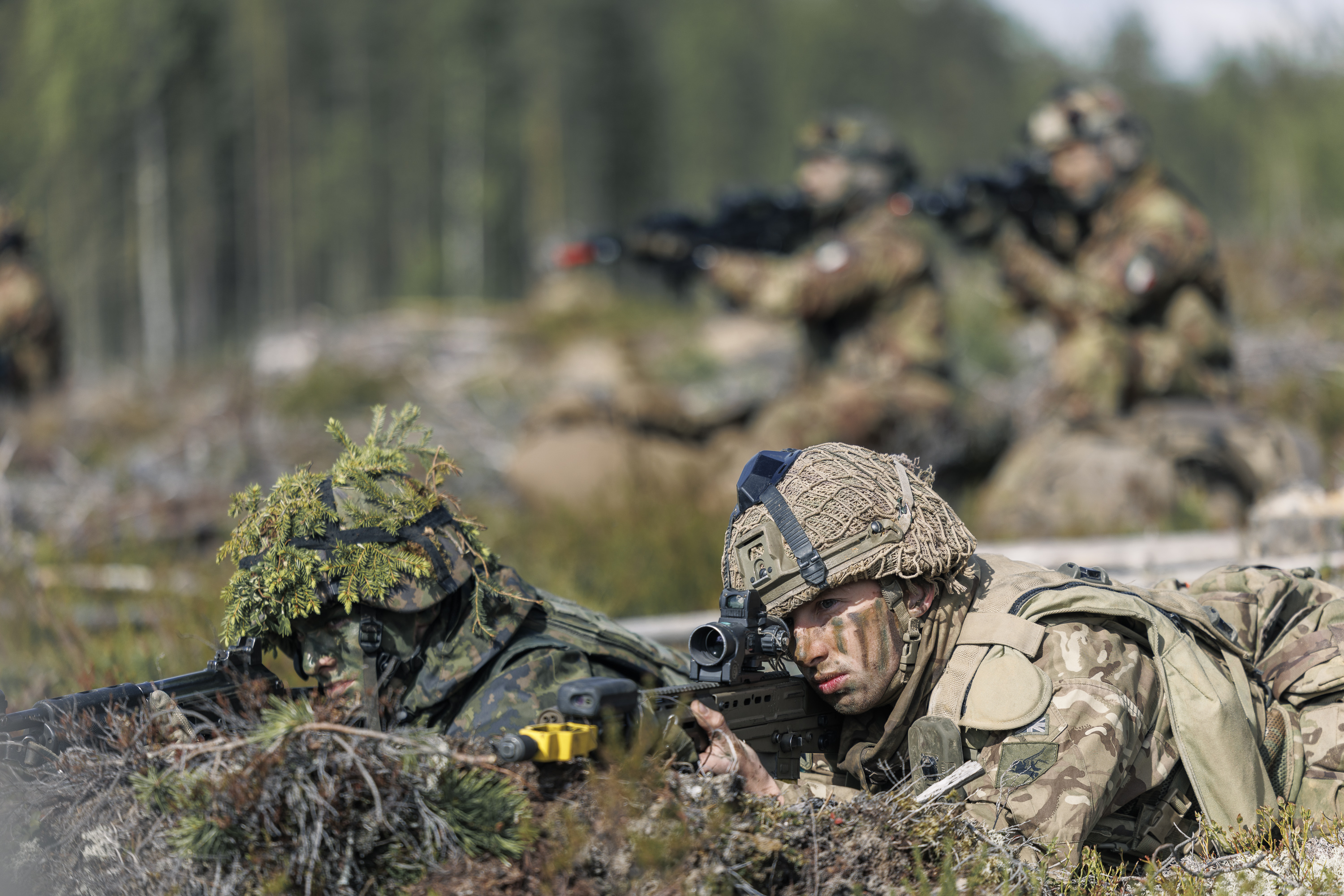Soldiers in camouflage uniforms lie prone on a forest floor, aiming rifles. The setting is a training exercise, conveying focus and readiness. Blurred trees in the background.
