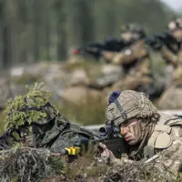 Soldiers in camouflage uniforms lie prone on a forest floor, aiming rifles. The setting is a training exercise, conveying focus and readiness. Blurred trees in the background.