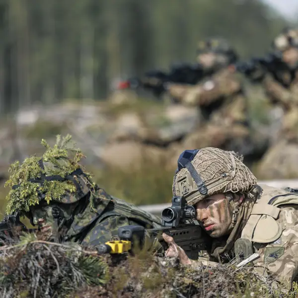 Soldiers in camouflage uniforms lie prone on a forest floor, aiming rifles. The setting is a training exercise, conveying focus and readiness. Blurred trees in the background.