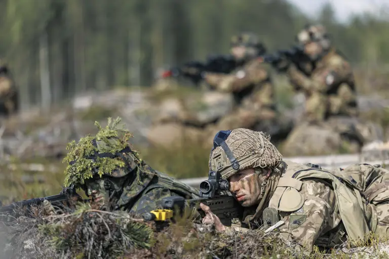 Soldiers in camouflage uniforms lie prone on a forest floor, aiming rifles. The setting is a training exercise, conveying focus and readiness. Blurred trees in the background.