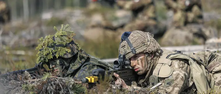 Soldiers in camouflage uniforms lie prone on a forest floor, aiming rifles. The setting is a training exercise, conveying focus and readiness. Blurred trees in the background.