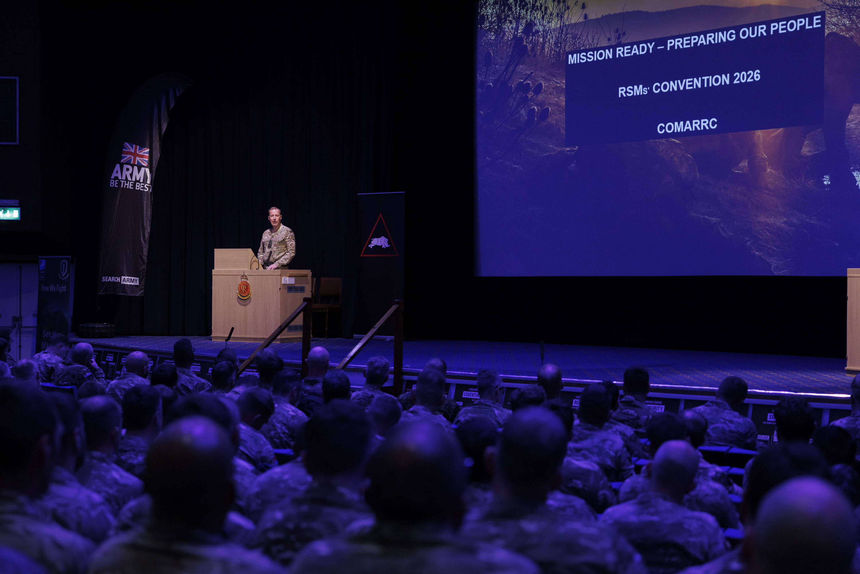 A military officer stands at a podium addressing uniformed soldiers seated in an auditorium. A screen reads 