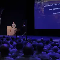 A military officer stands at a podium addressing uniformed soldiers seated in an auditorium. A screen reads